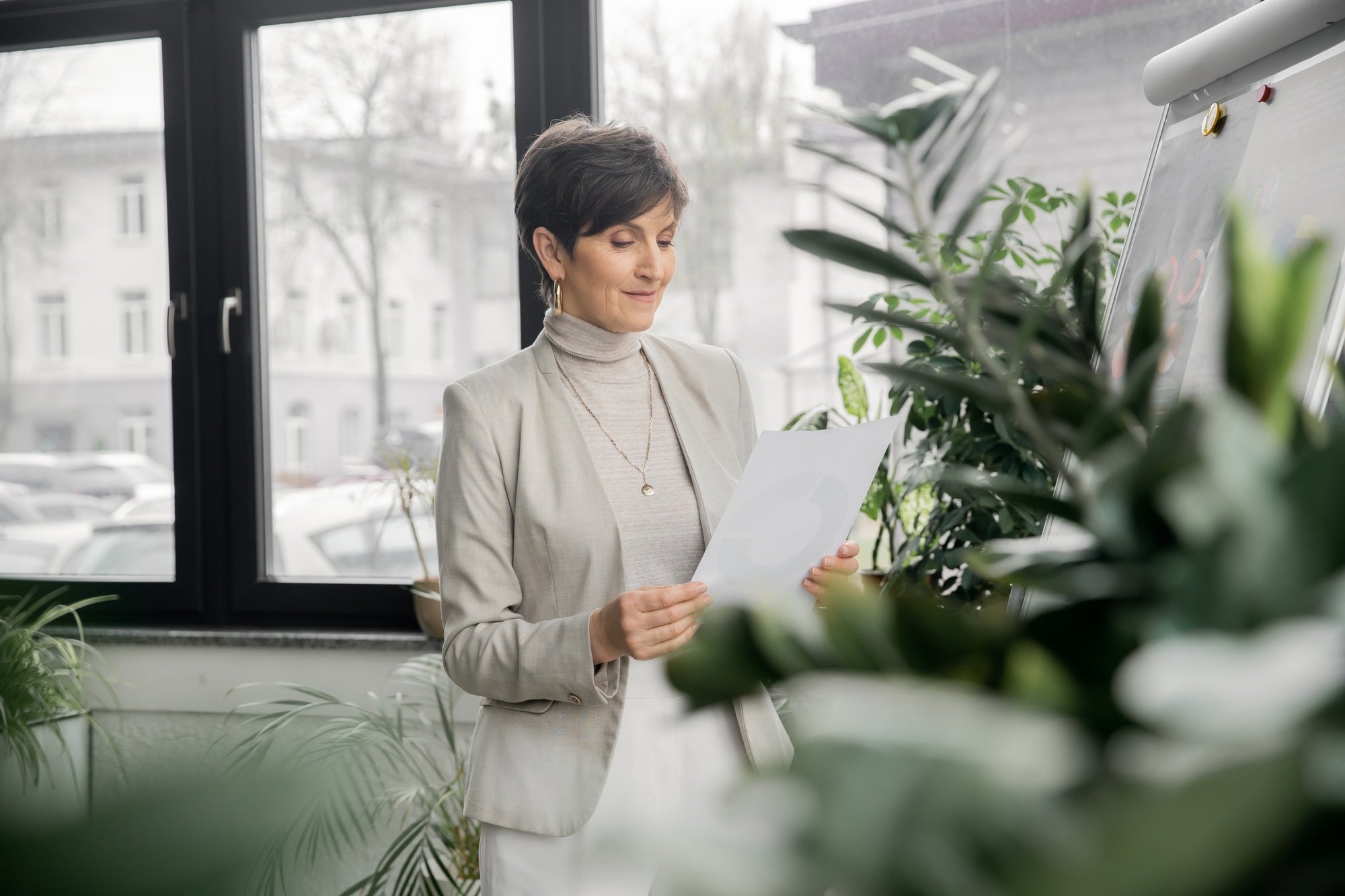 positive middle aged businesswoman looking at document, working in modern office, project planning