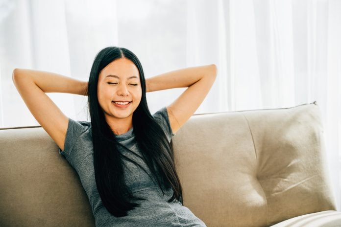Relaxed woman on sofa hands behind head enjoys serene moment