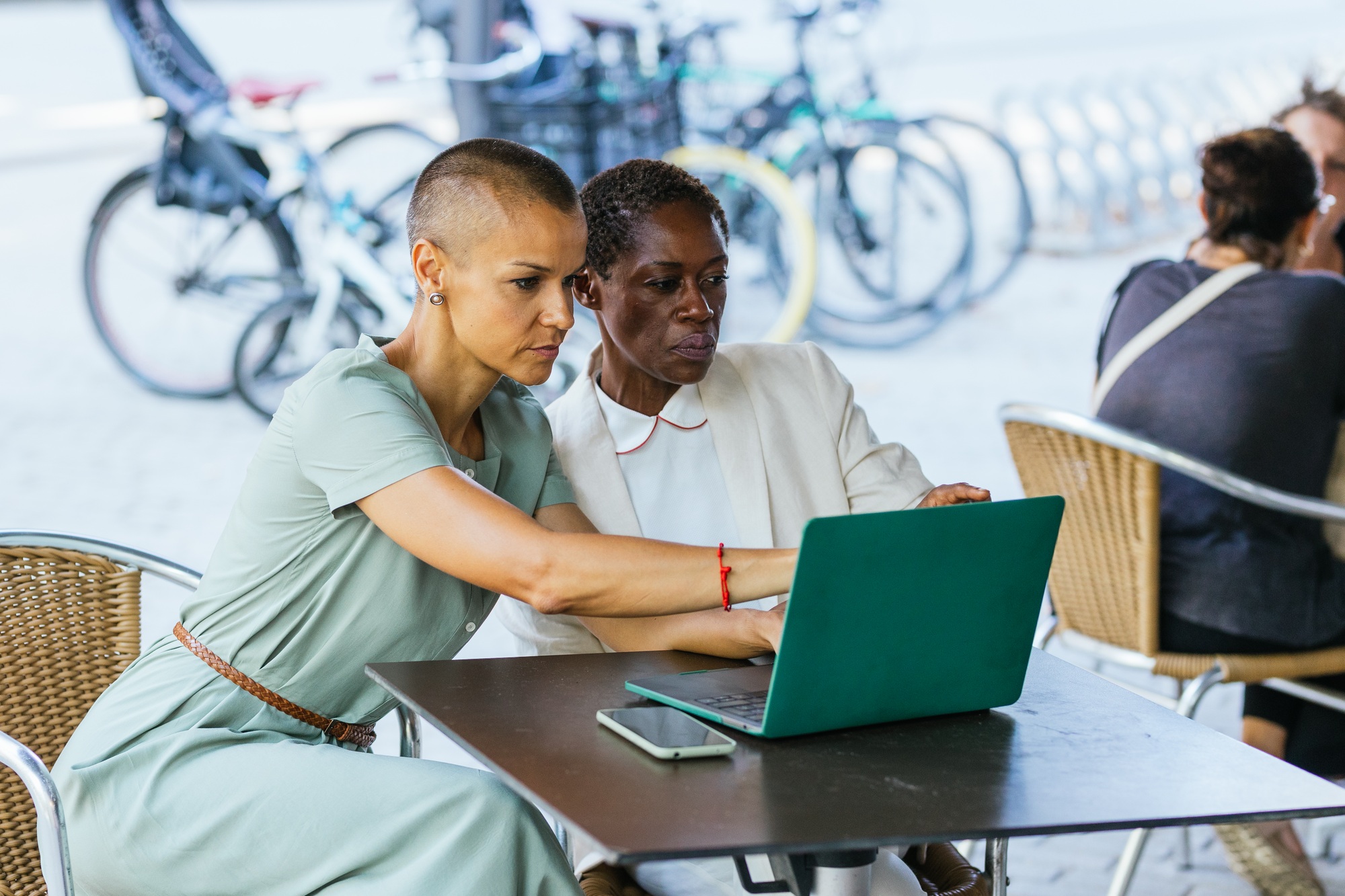 Two businesswomen on the terrace of a coffee shop are discussing a job on the laptop and have a