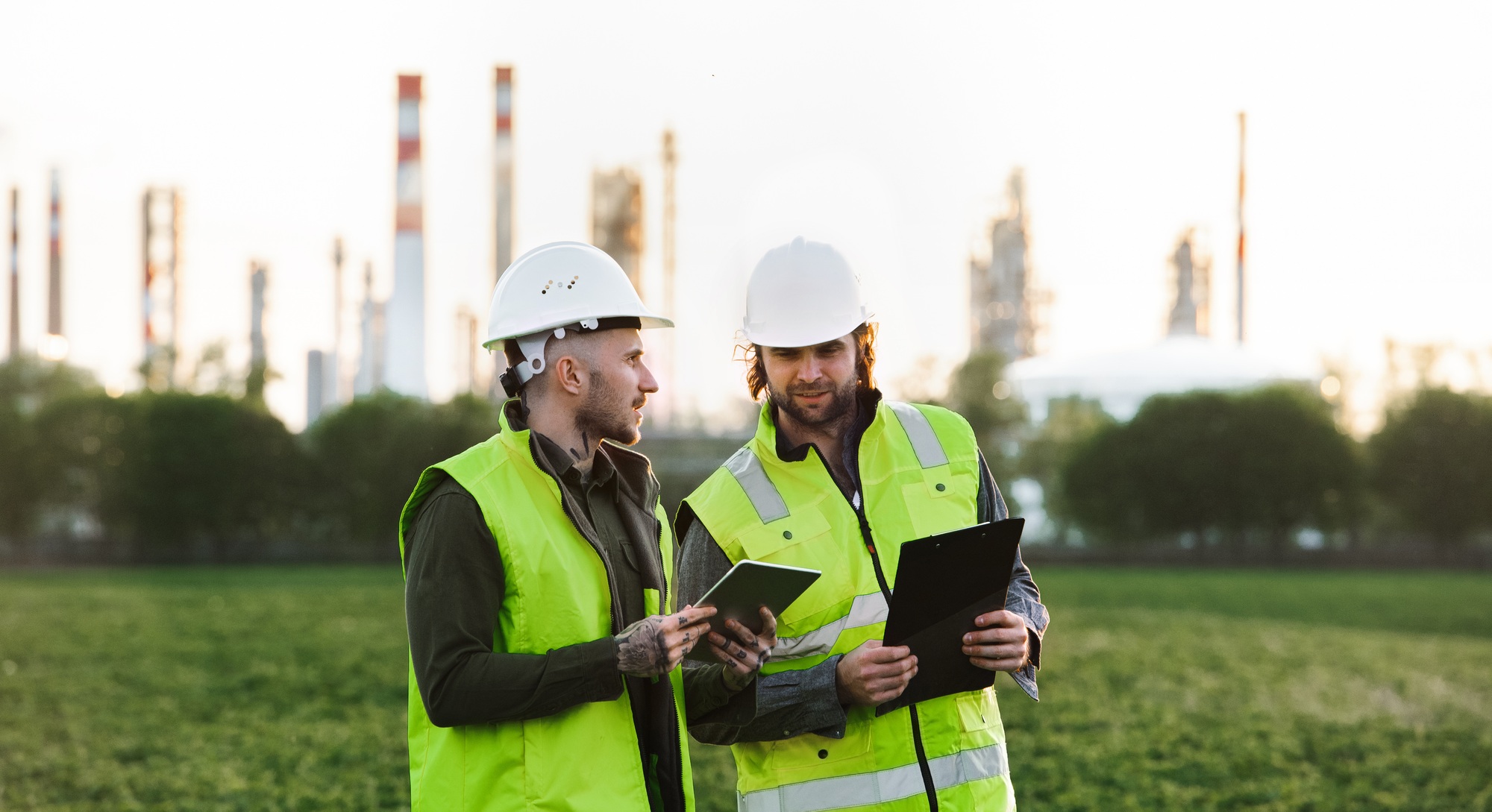 Two young engineers with tablet standing outdoors by oil refinery, discussing issues