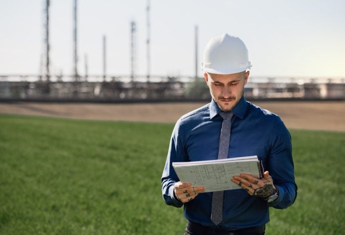 Young engineer with hard hat and tablet standing outdoors by oil refinery