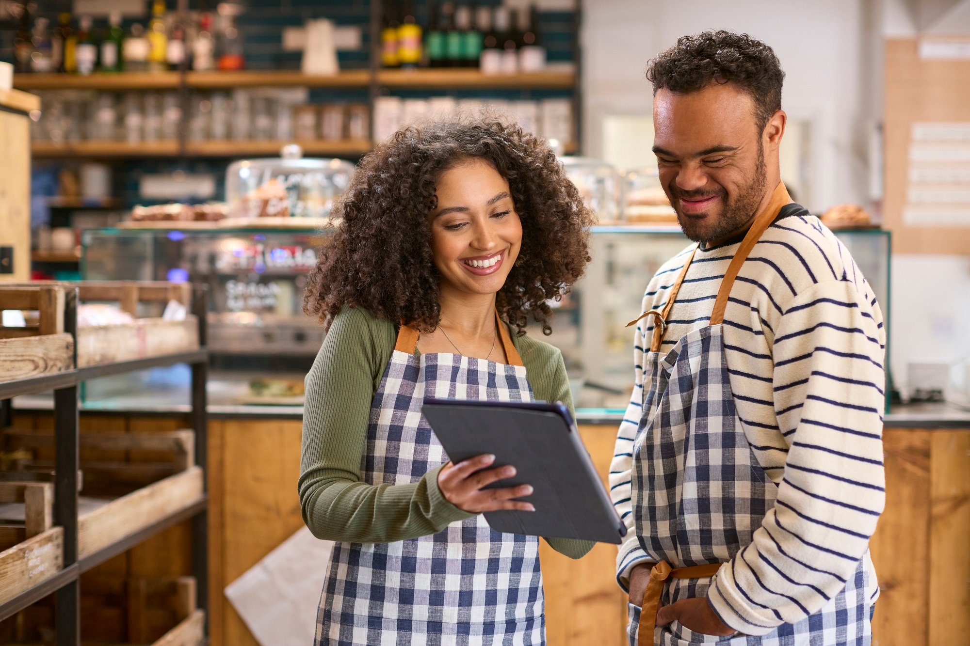 Woman Using Digital Tablet Working In Food Shop Training Man With Down Syndrome