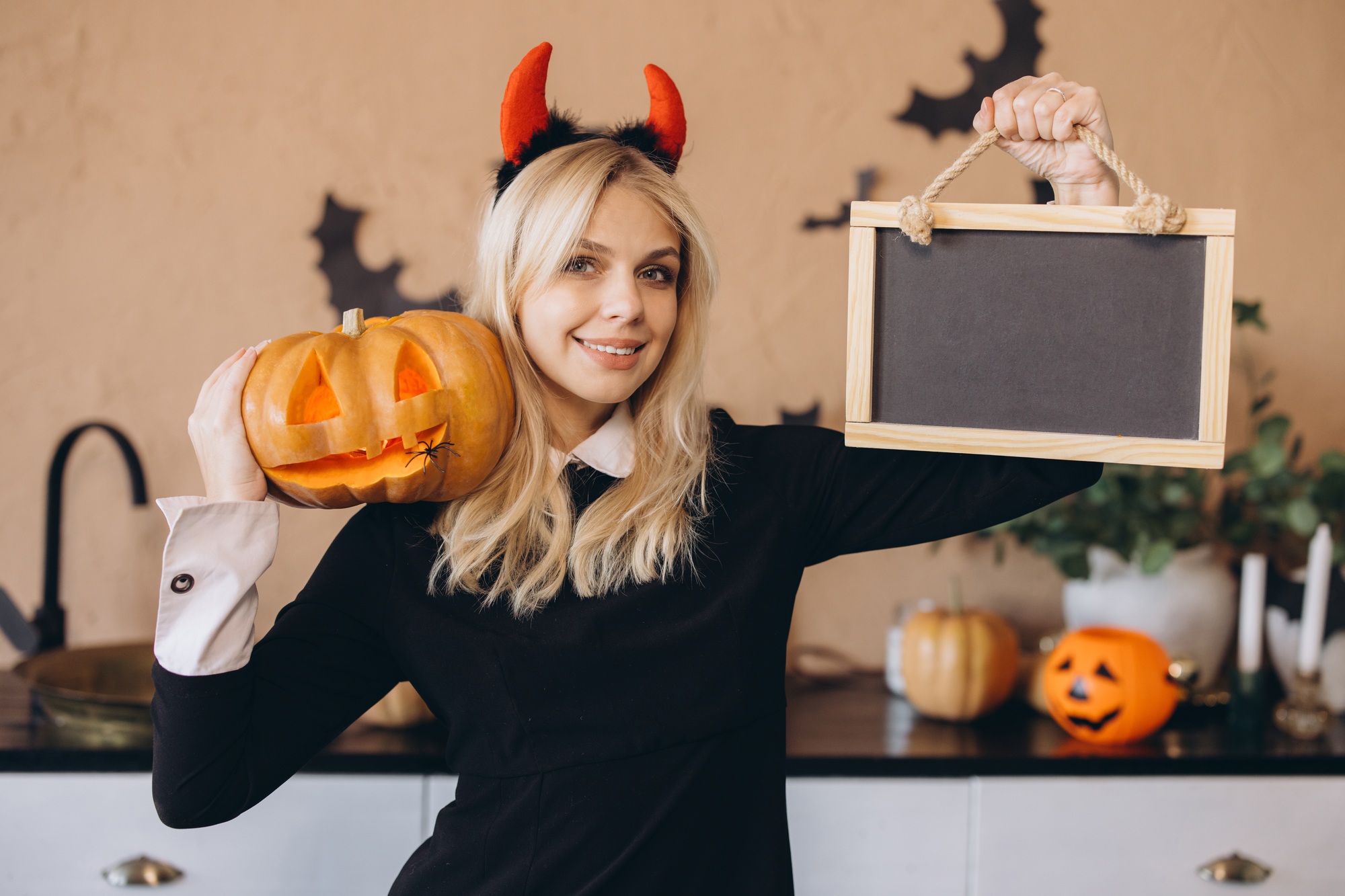 Woman wearing devil costume holding jack o' lantern and blank blackboard celebrating Halloween