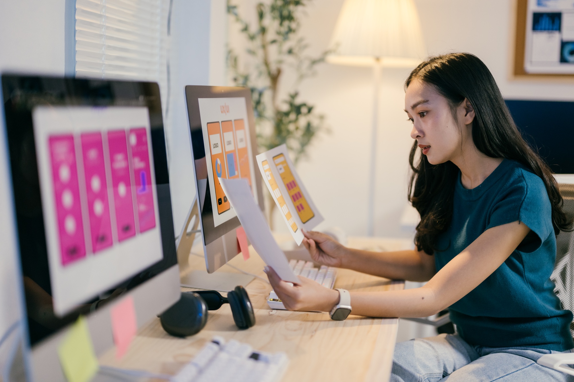 Young woman working as a web designer is comparing two website mockups while working at her desk. Sh
