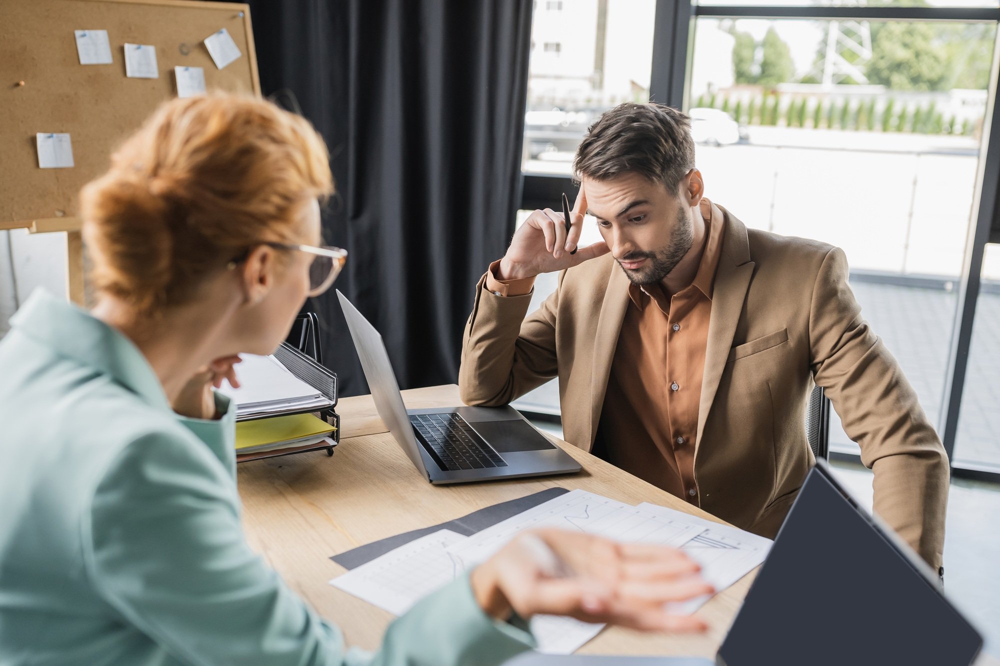 discouraged manager looking at papers near blurred businesswoman gesturing during conflict in office