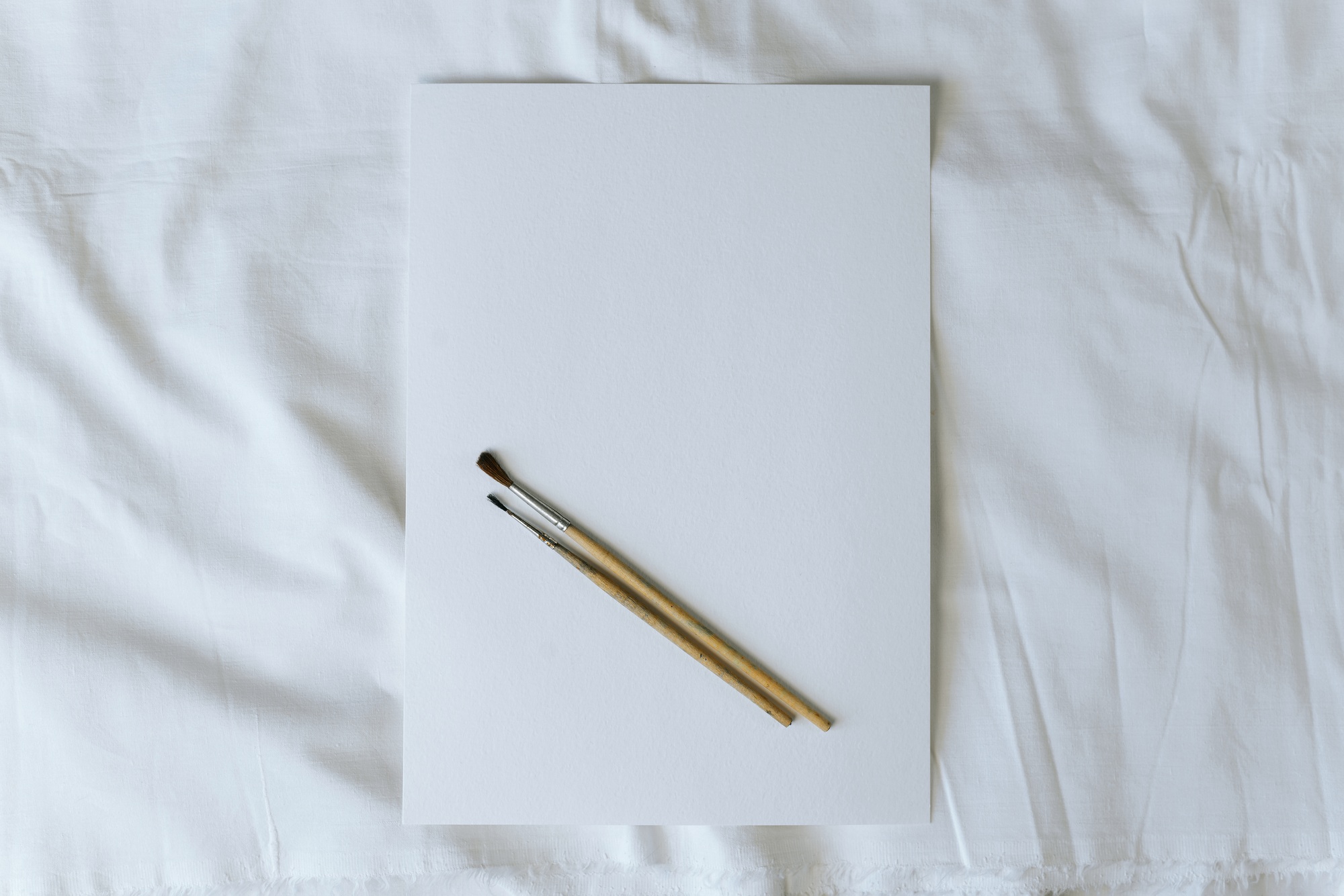 Empty sheet of watercolor paper and two paint brushes lying on a white cloth, flat lay