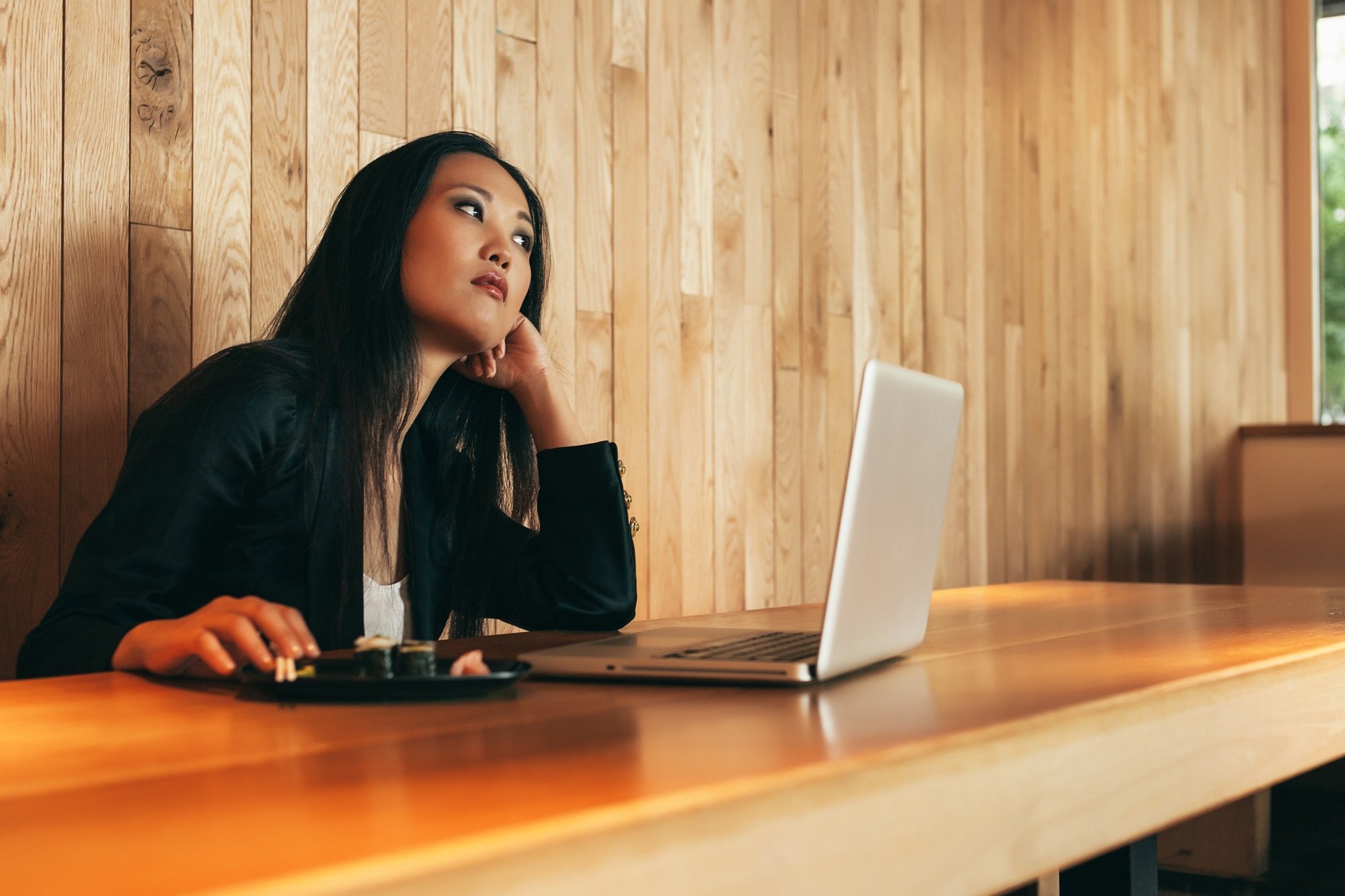 Ethnic businesswoman eating sushi and working on laptop in cafe