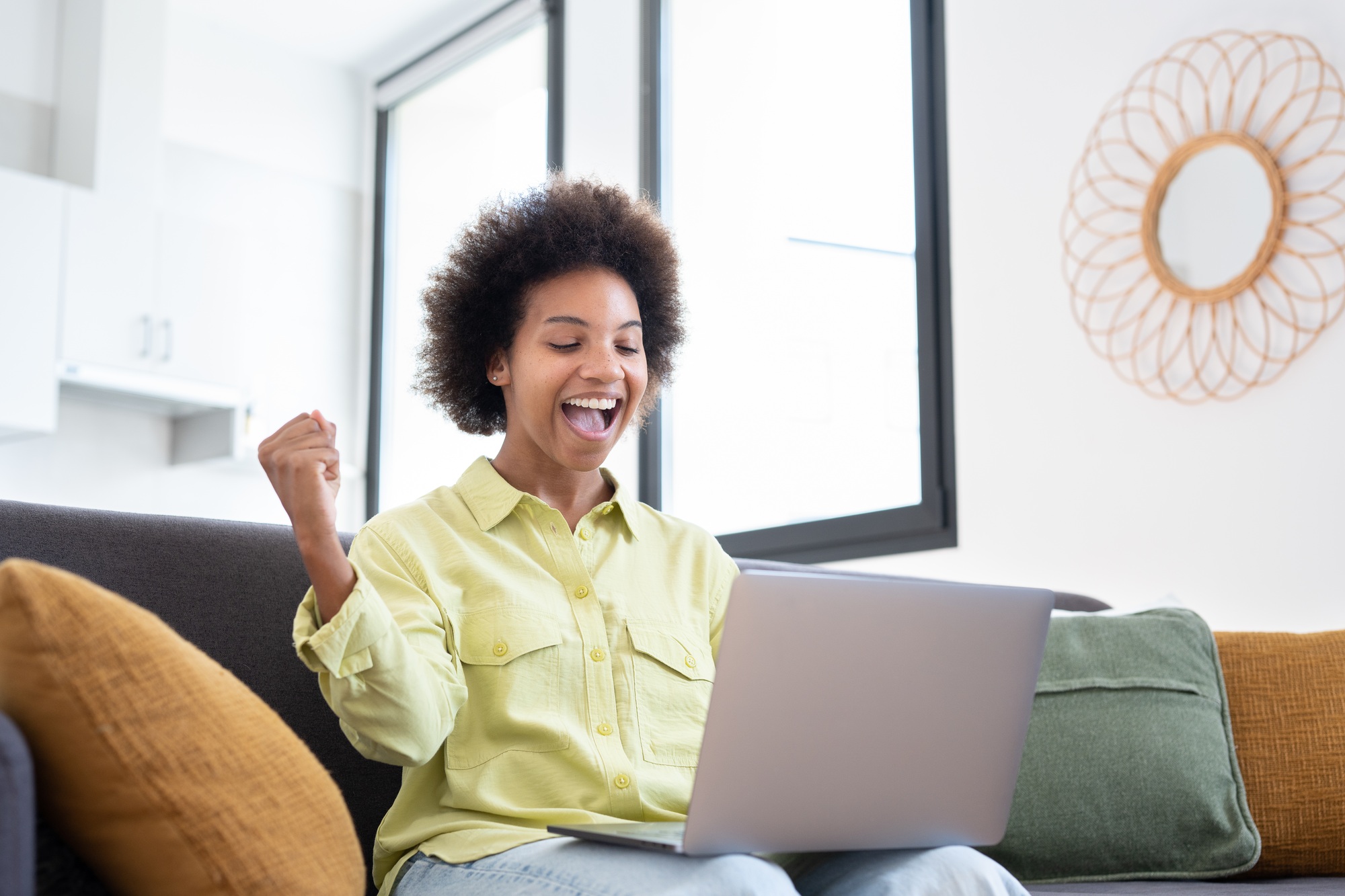 Excited cheerful young Black woman using laptop computer on sofa at home, getting good news