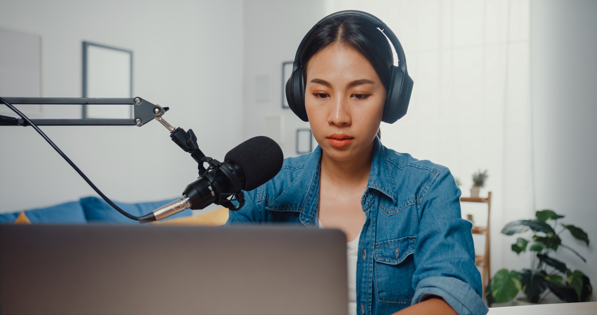 Girl record a podcast on laptop computer with headphones and microphone talk at living room.
