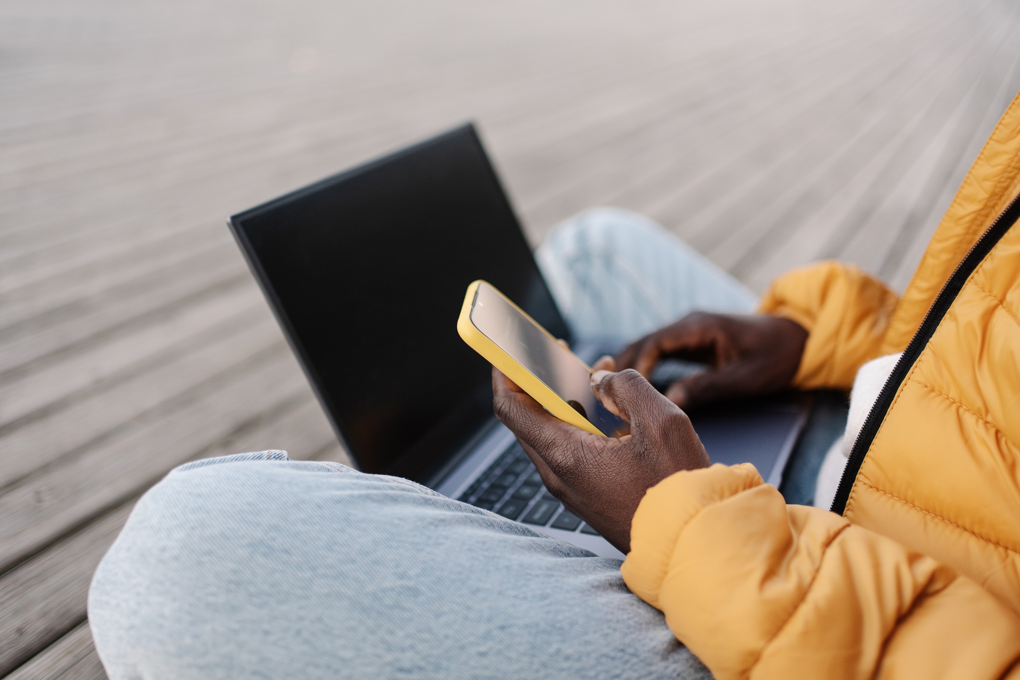 Hand of African American man working on laptop using yellow cell phone to verify account