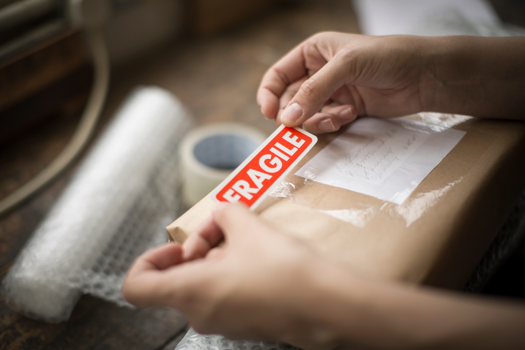 Hands holding a red Fragile sticker to stick it on a brown paper package on a work bench.