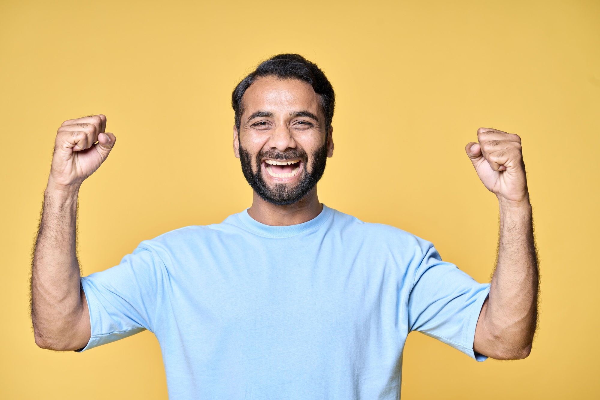 Happy excited indian man raising fists isolated on yellow background.