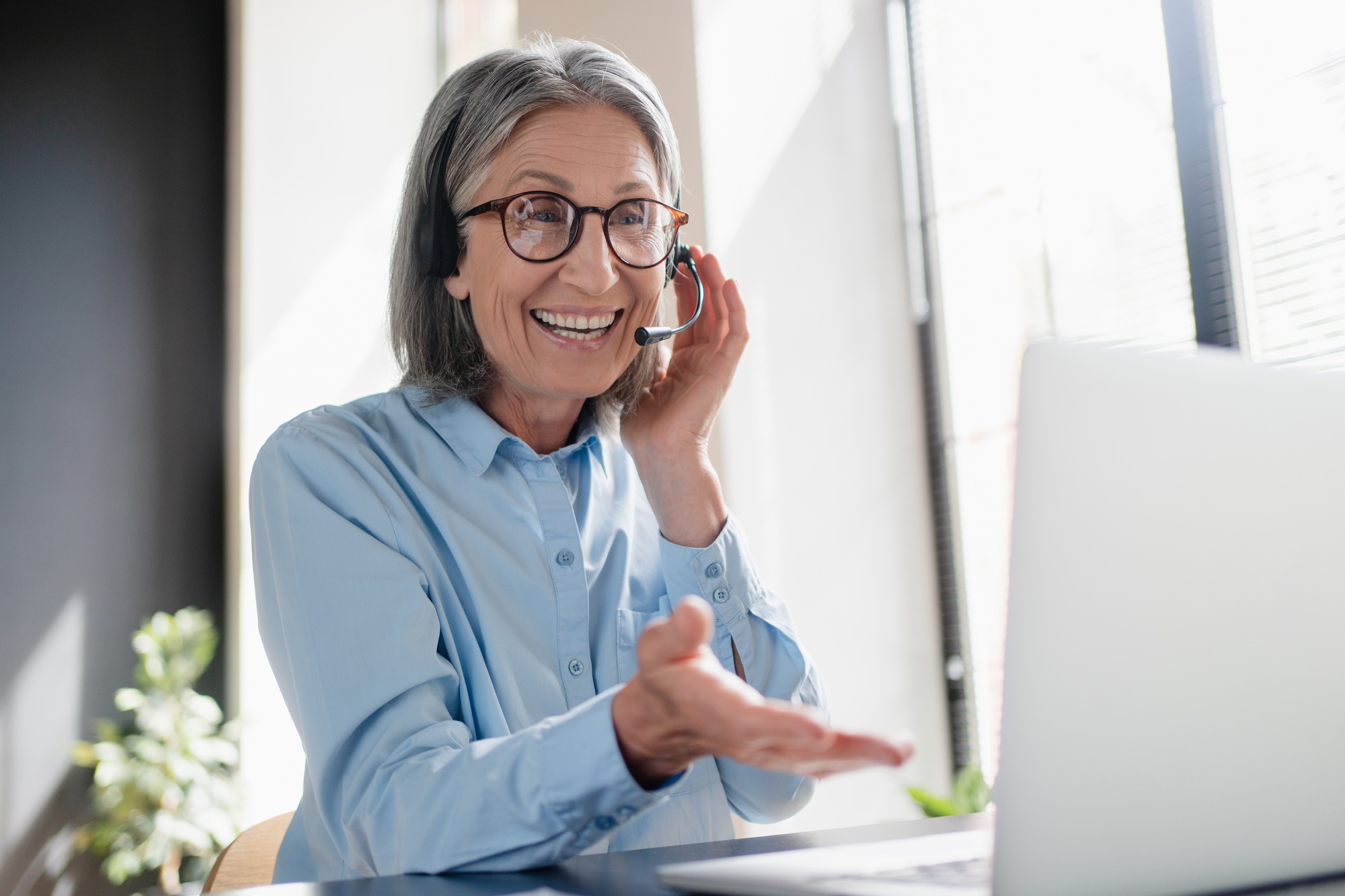 Happy mature woman call contact center agent wearing headset working in business office using laptop