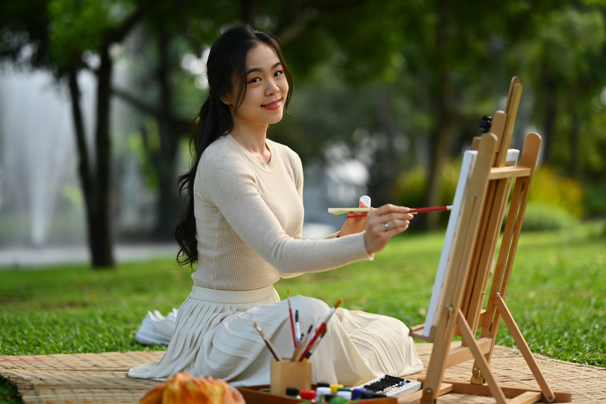 Image of smiling young woman painting on canvas enjoying free time at green park