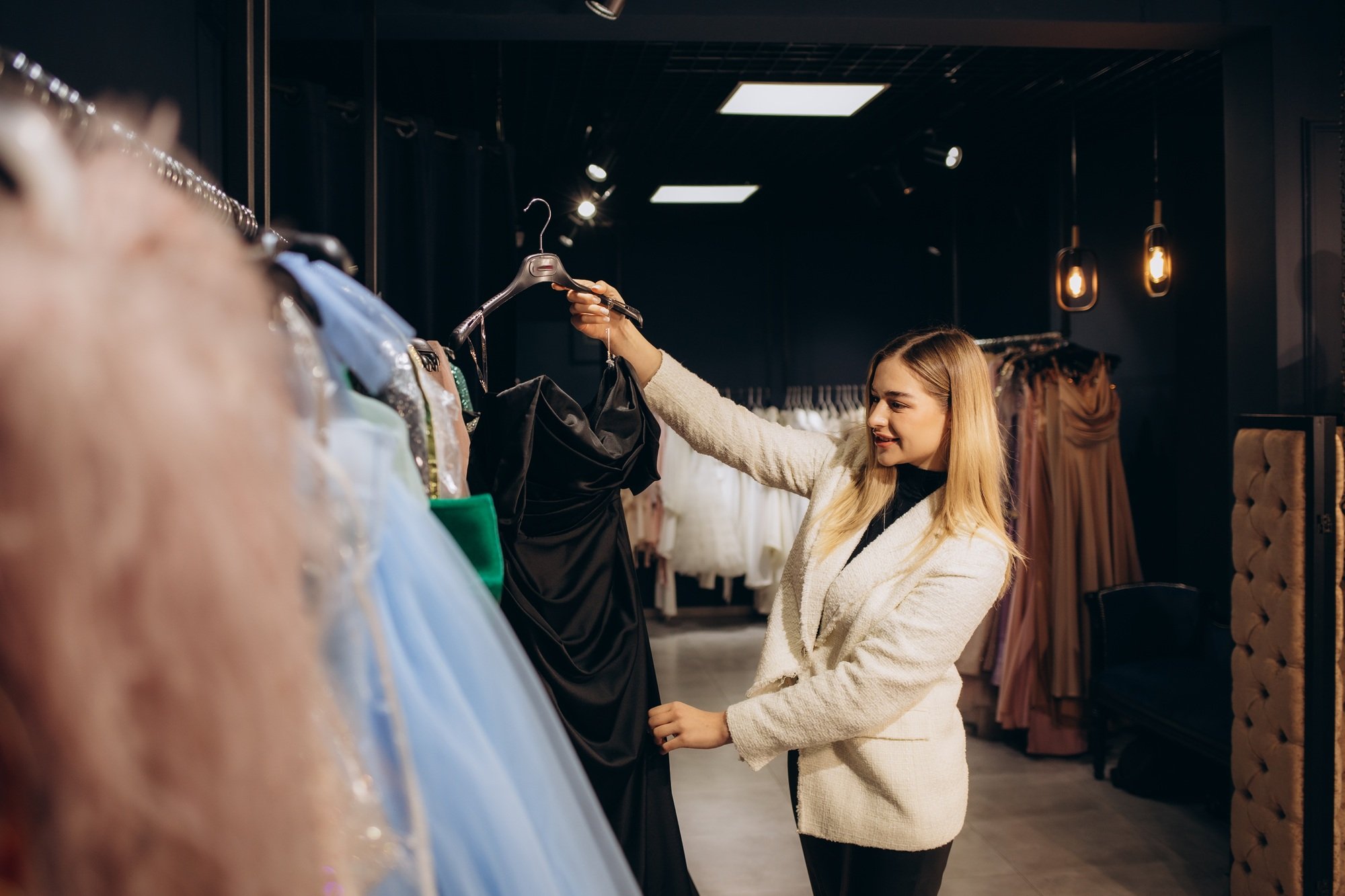 Image of young lady standing in clothes shop indoors choosing dresses. Looking aside.