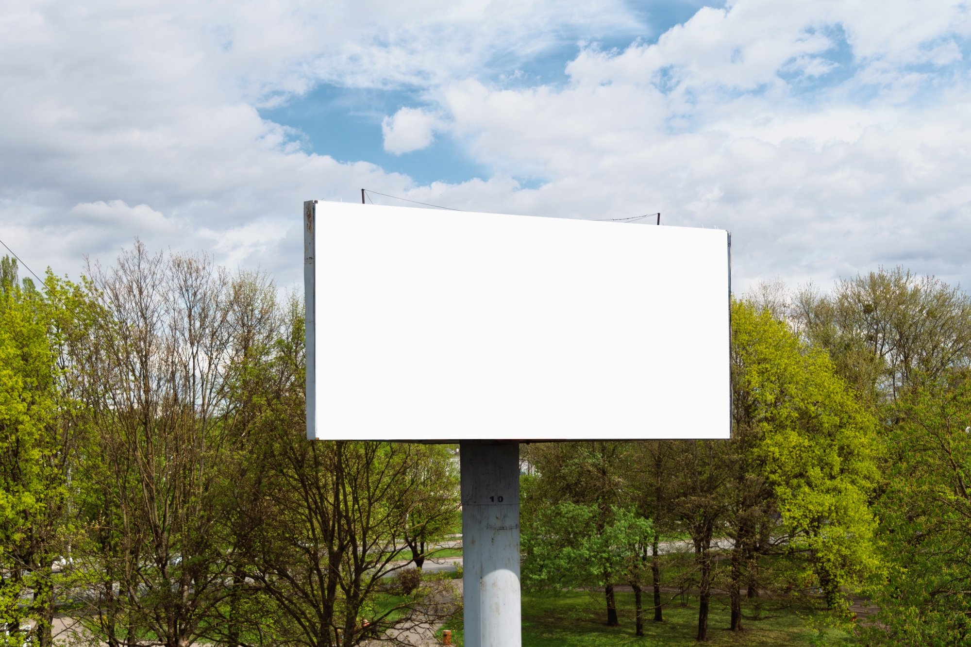 Large blank billboard stands against a backdrop of green trees and blue sky