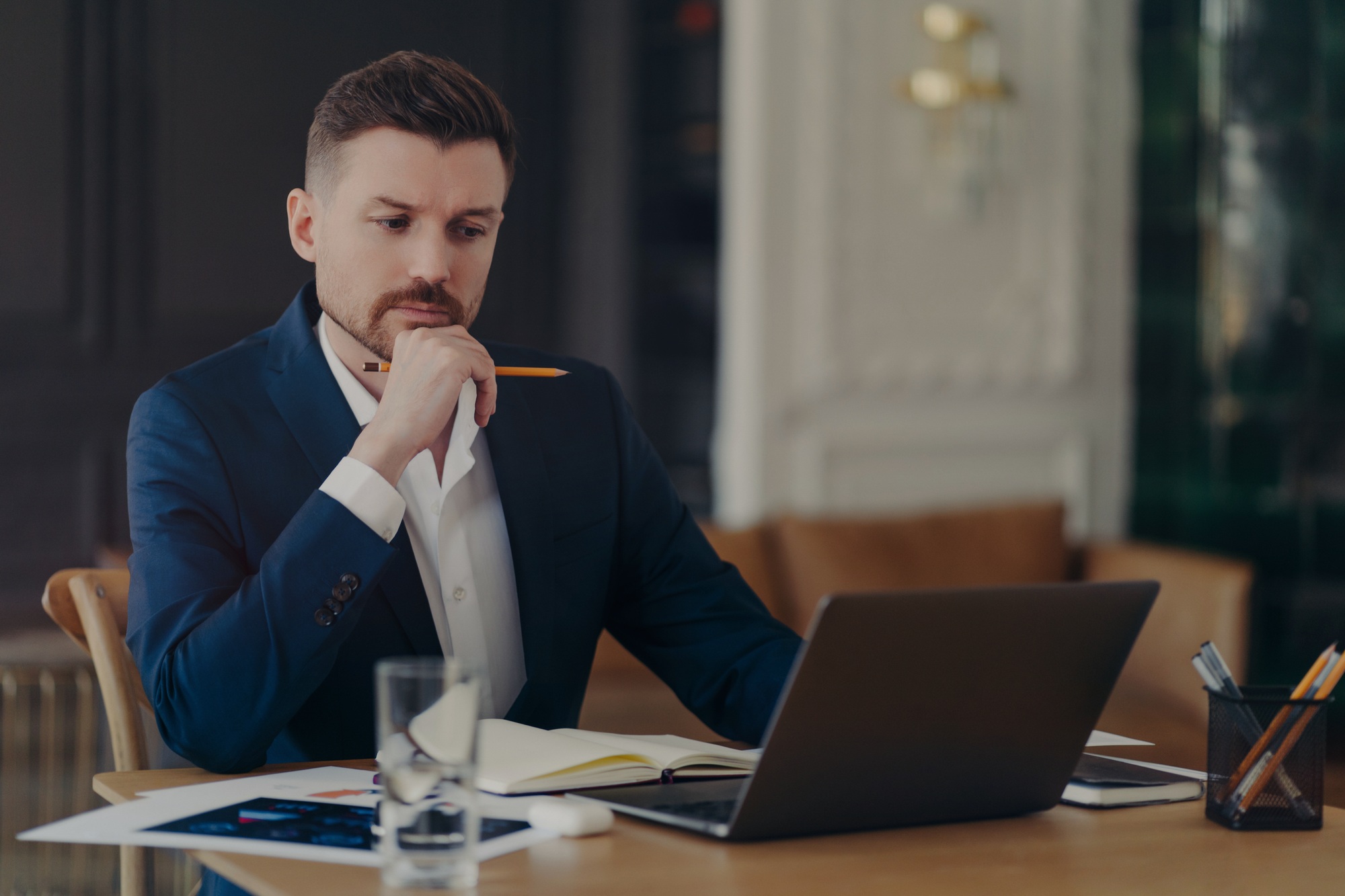 Male entrepreneur holds chin makes notes in diary works on laptop computer makes money online