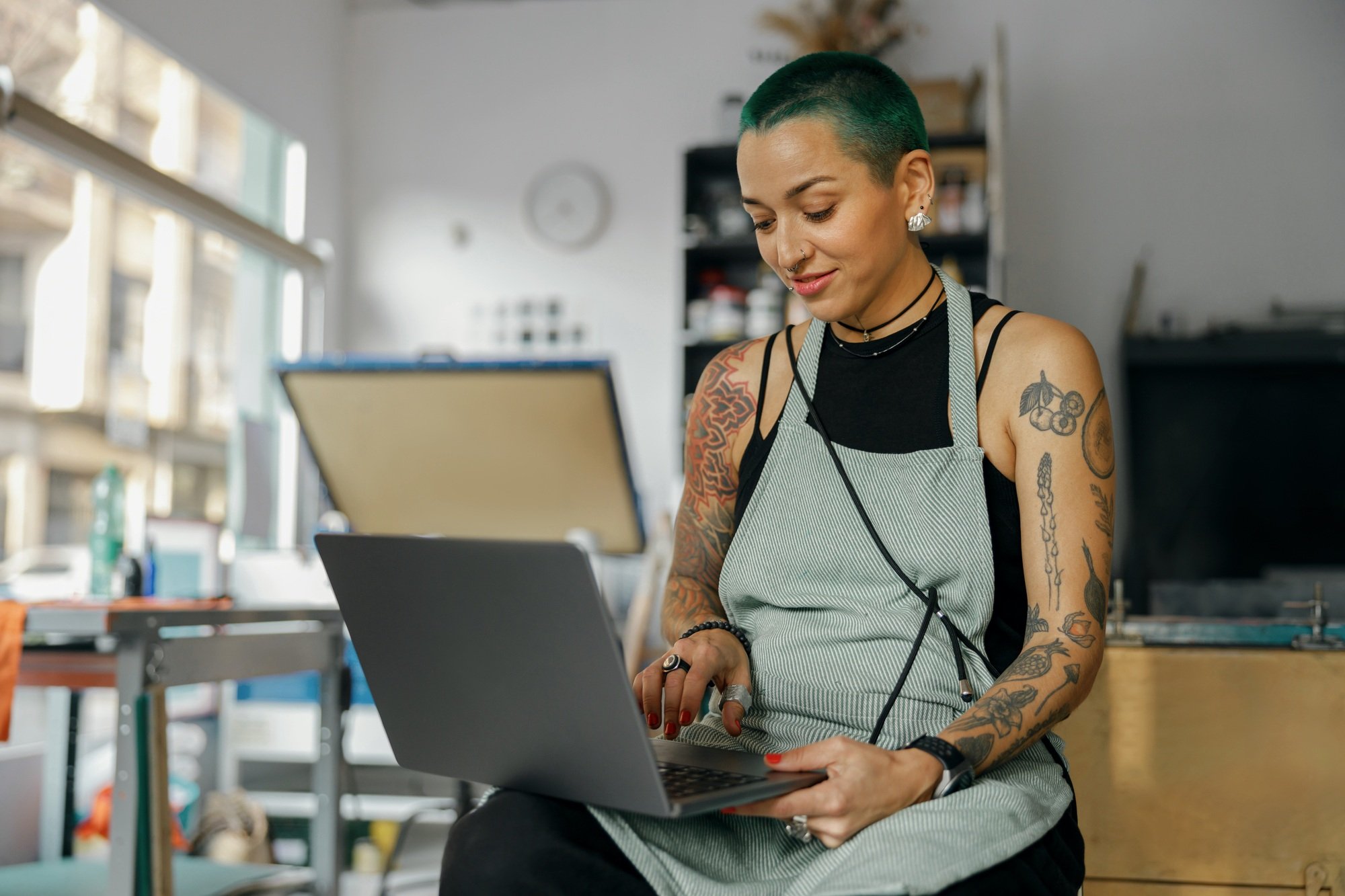 Positive female designer sitting with laptop on background of print machine in workshop
