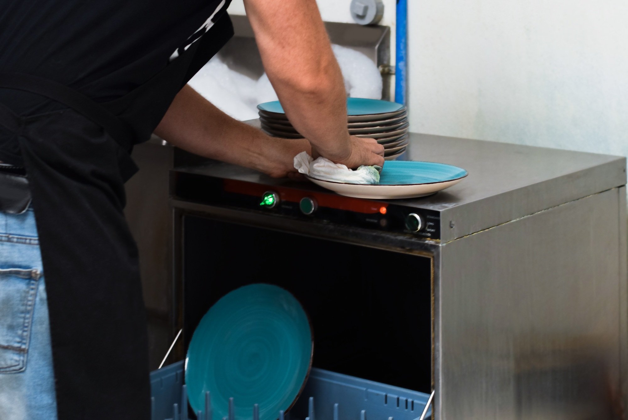 Restaurant kitchen employee taking out dishes from dishwasher and wiping with clean towel