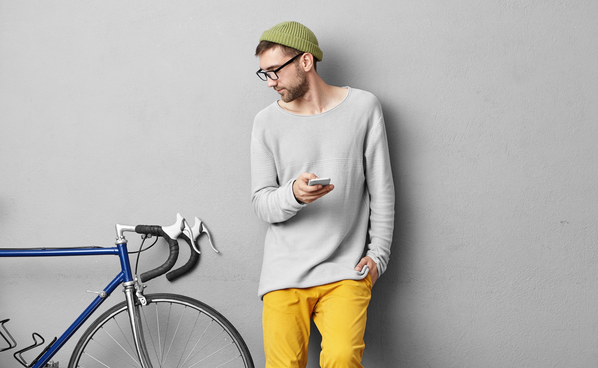 Serious unshaven young man in trendy wear posing at concrete wall and looking at his fixed gear bike