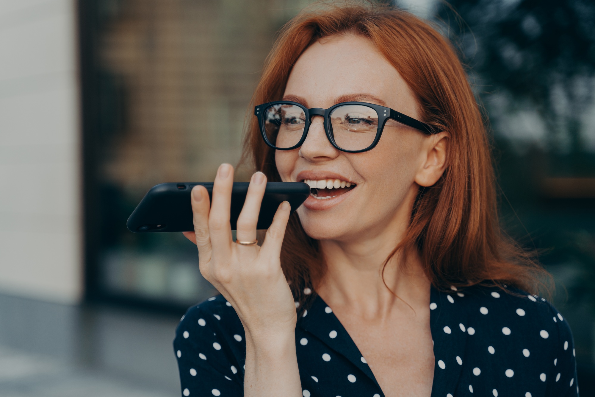 Smiling red-haired business woman using virtual digital voice assistant while standing outdoors