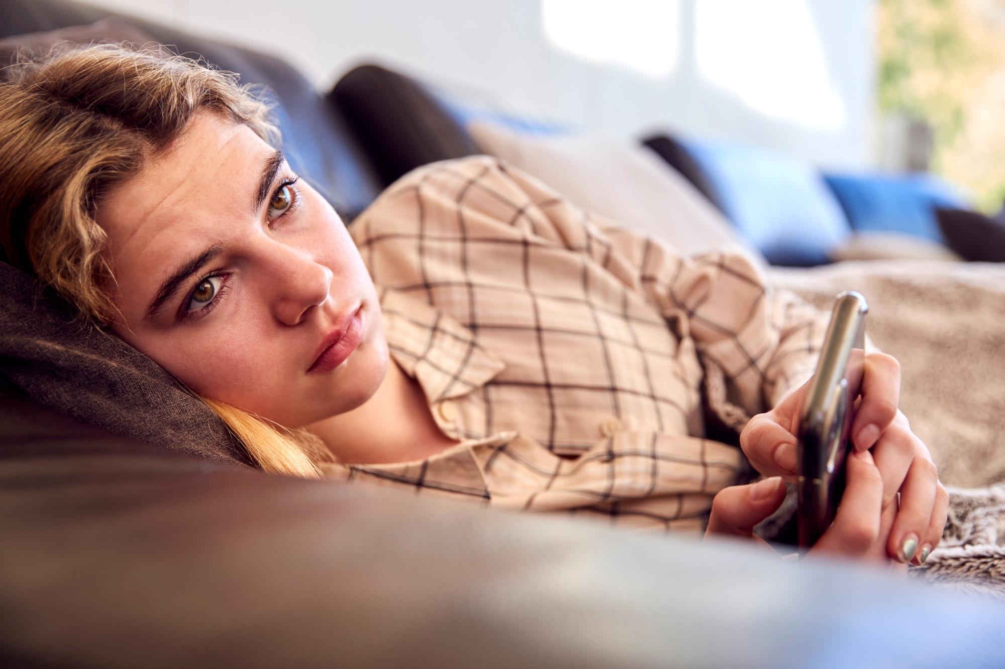 Unhappy Teenage Girl Lying On Sofa With Mobile Phone Being Bullied Online At Home