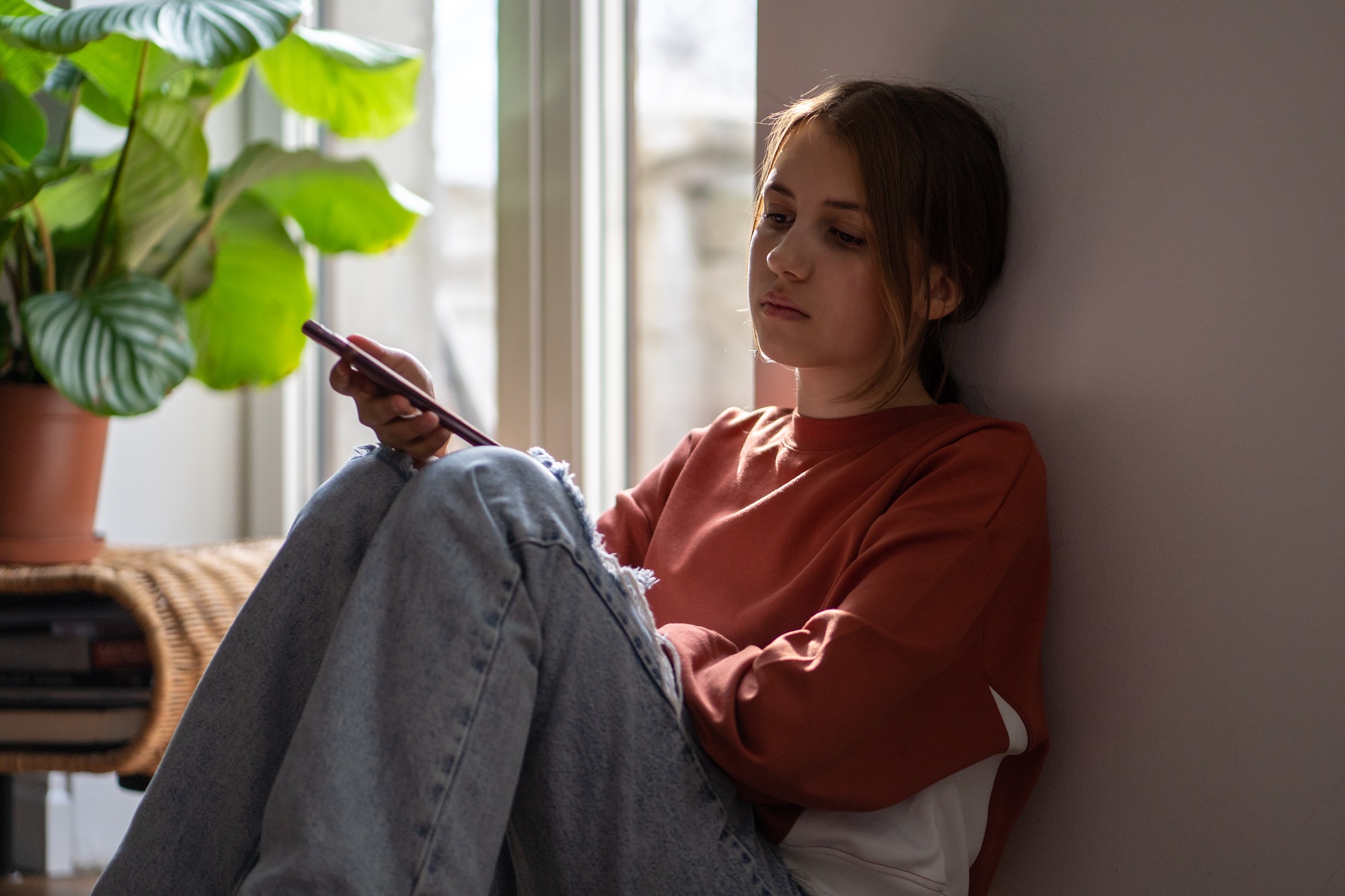 Upset anxious teen girl reads message in smartphone sits on floor at home near window.