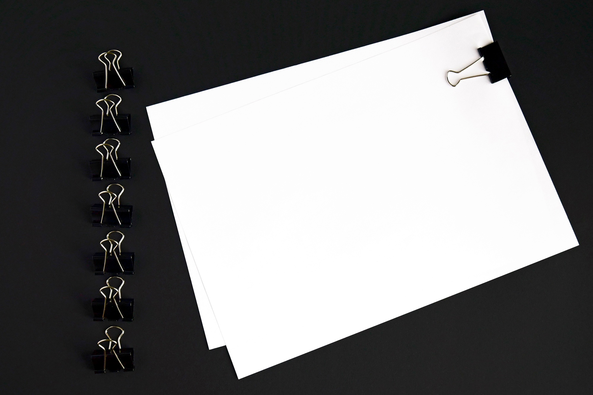 White sheets and binder clip on black background, top view, mockup.