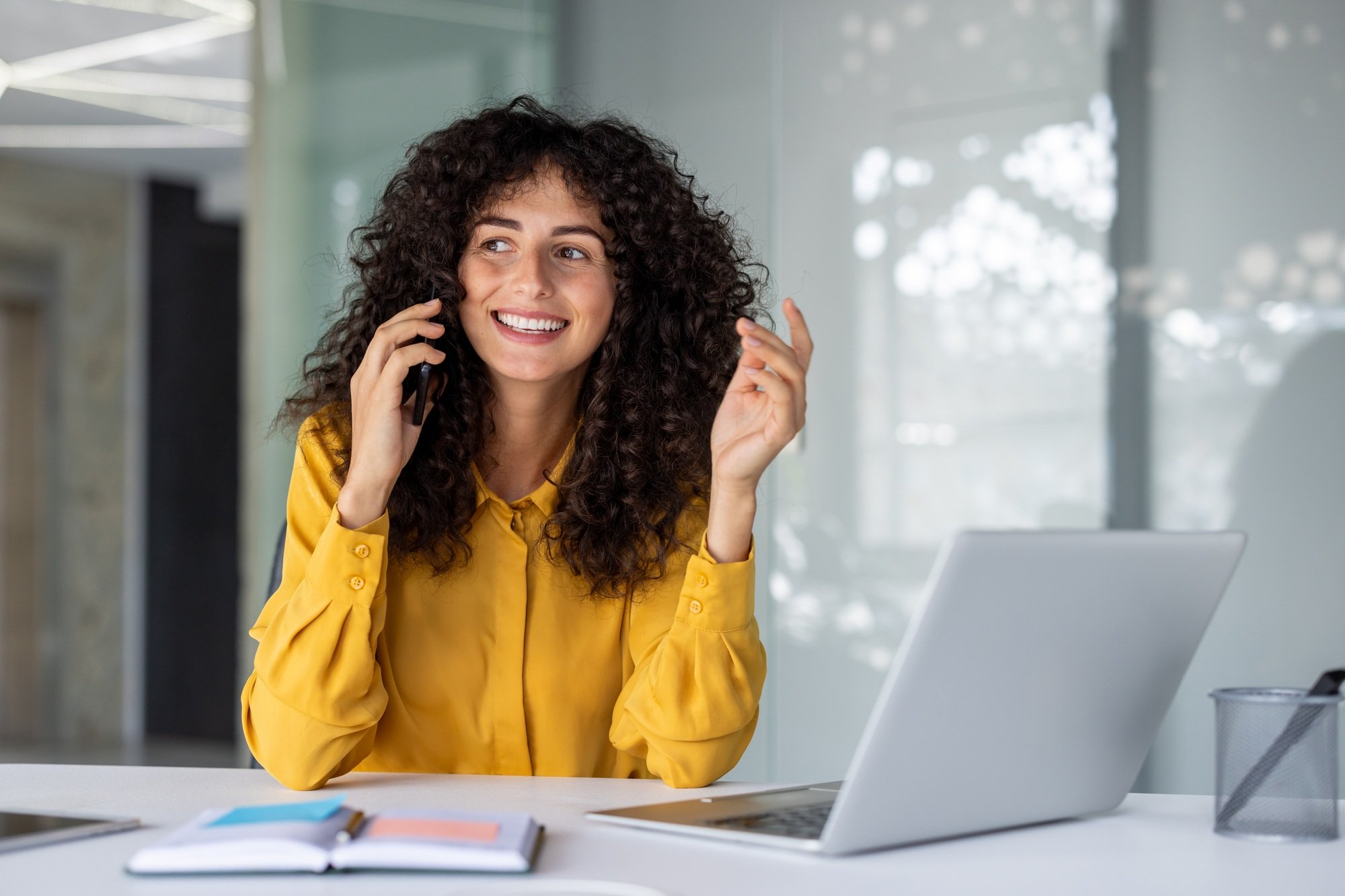 Woman in bright yellow blouse happily speaking on smartphone at modern workspace with laptop and