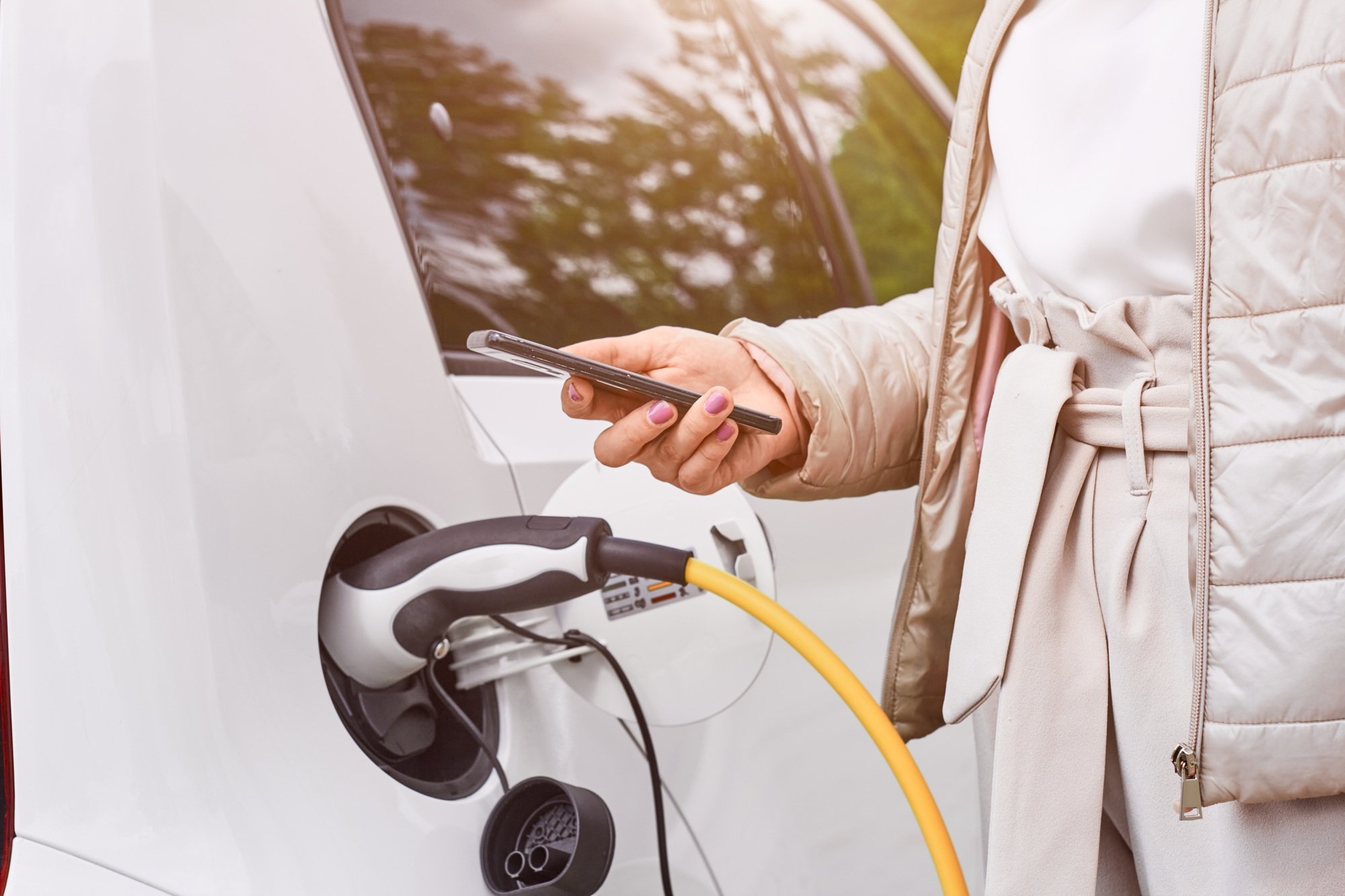Woman standing near charging electric car with a mobile phone in her hands. Car sharing or rental