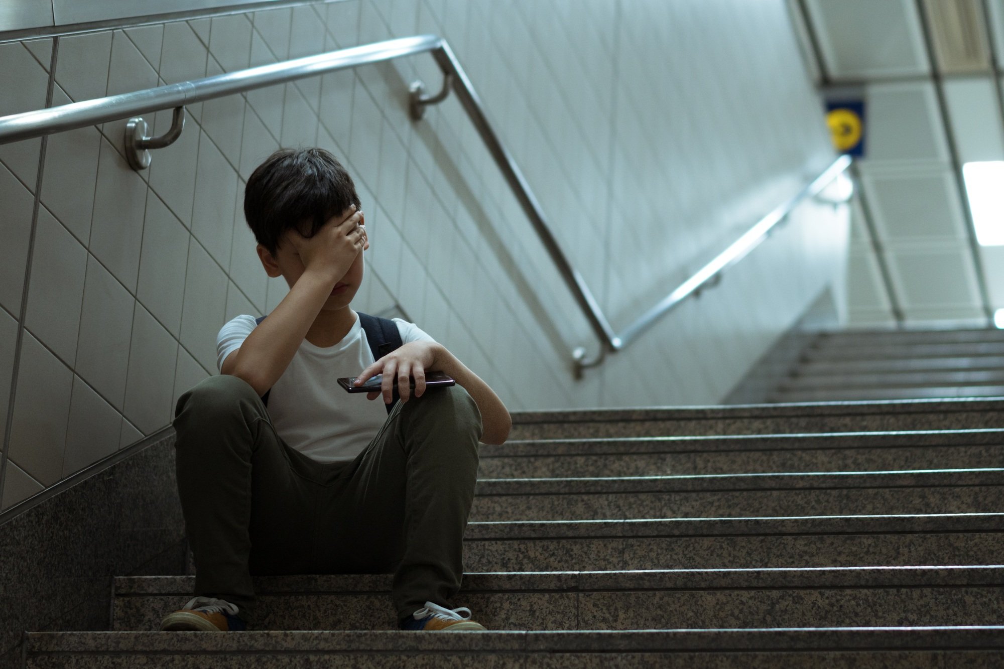 Young Asian boy sit on stairs alone, hold smartphone and cover his face in despair and frustration