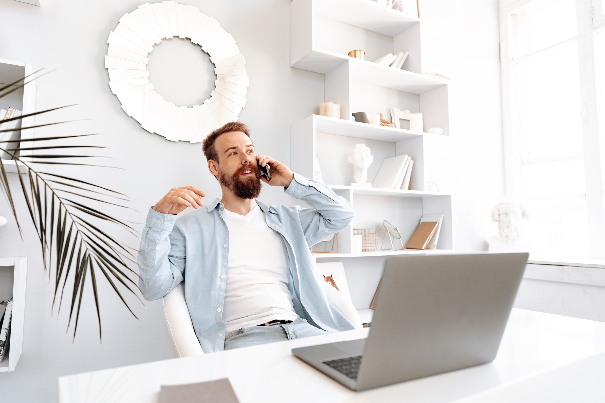 Young bearded businessman working from home sitting at the desk and talking on mobile phone