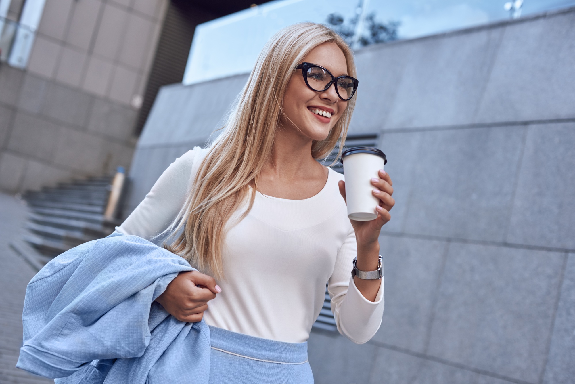 Young pretty spectacled businesswoman smiling and walking near business building with coffe