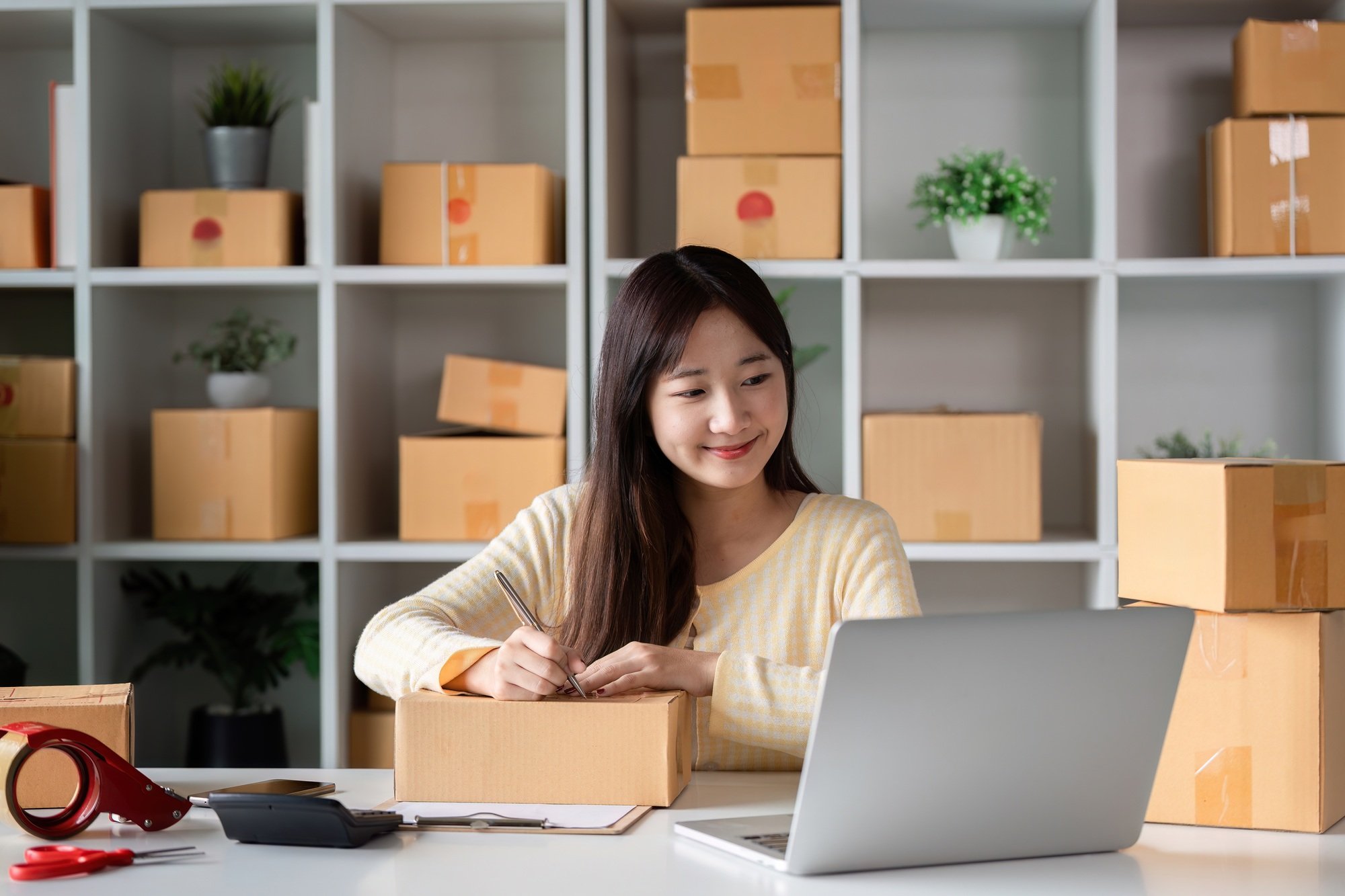 Young Woman Entrepreneur Checking Product Orders Online in a Modern Home Office with Shipping Boxes