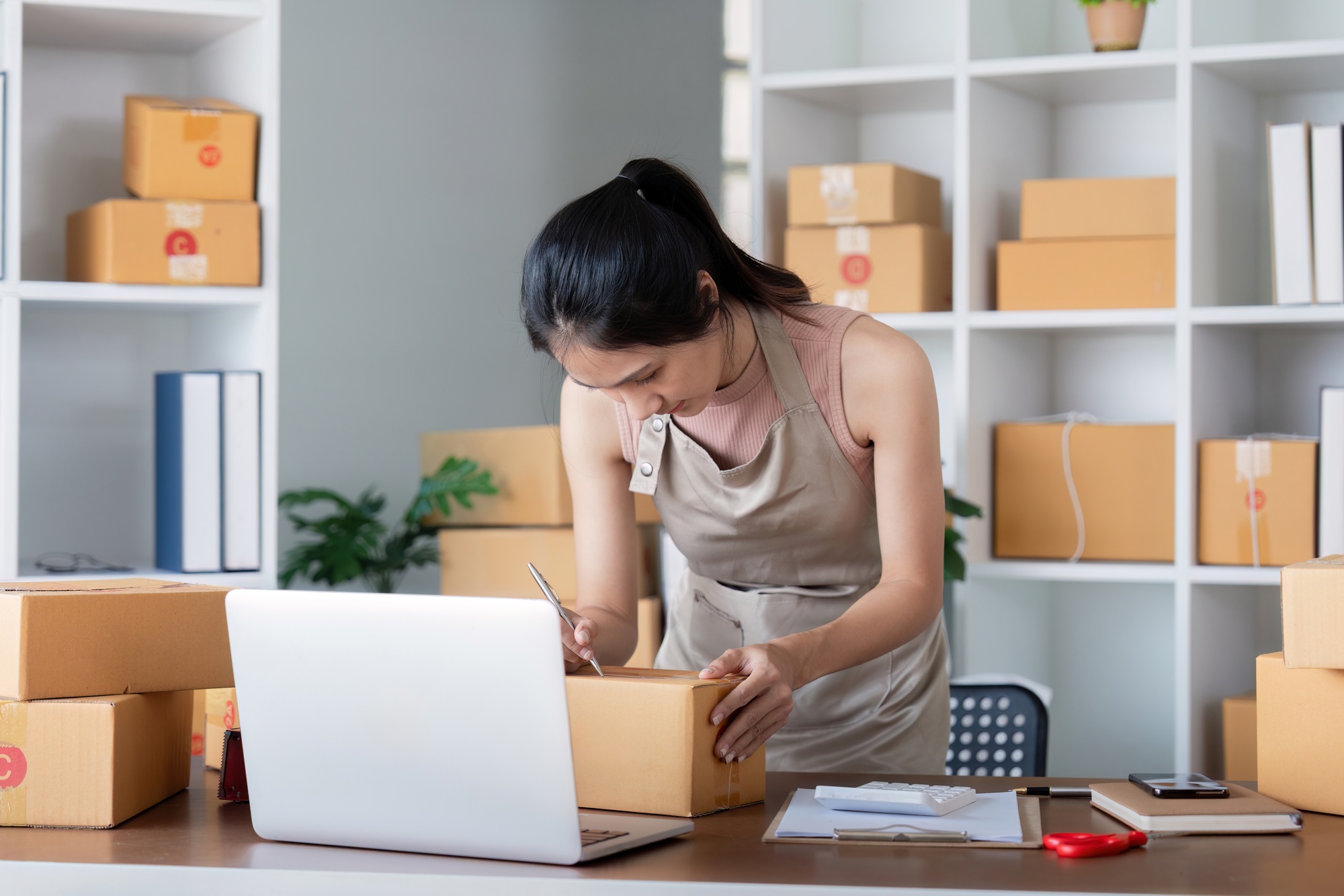 Young Woman Preparing Packages for Shipping in a Home Office with Laptop and Cardboard Boxes